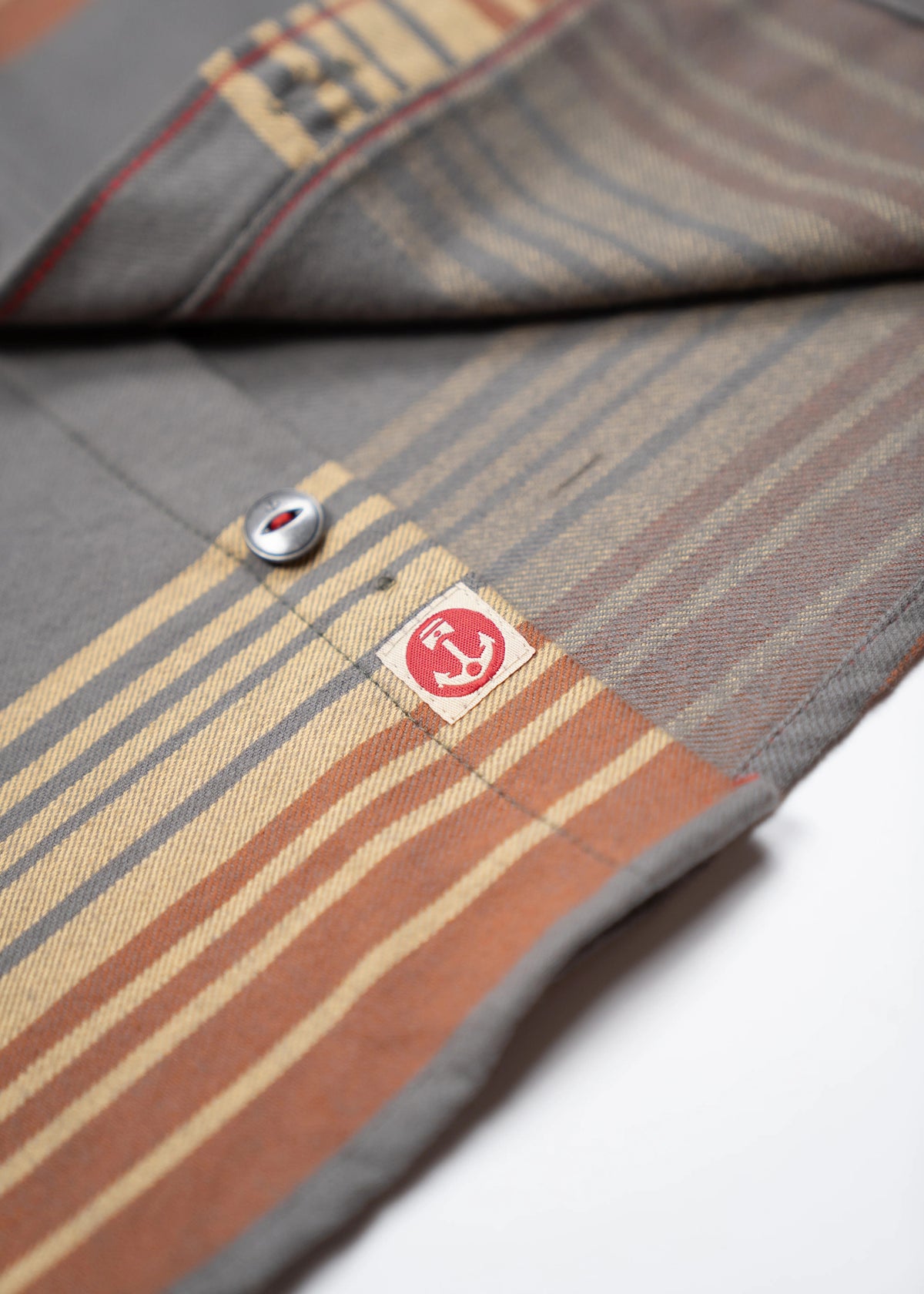 Close-up of a striped fabric with a button and red logo tag, featuring shades of gray, beige, and brown in a diagonal pattern.