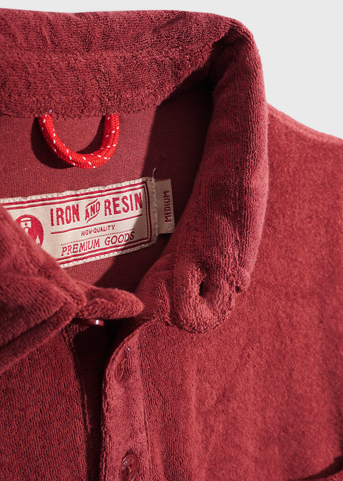Close-up of a red terry cloth shirt with a buttoned collar, featuring an Iron and Resin label marked "Premium Goods" and "Medium."
