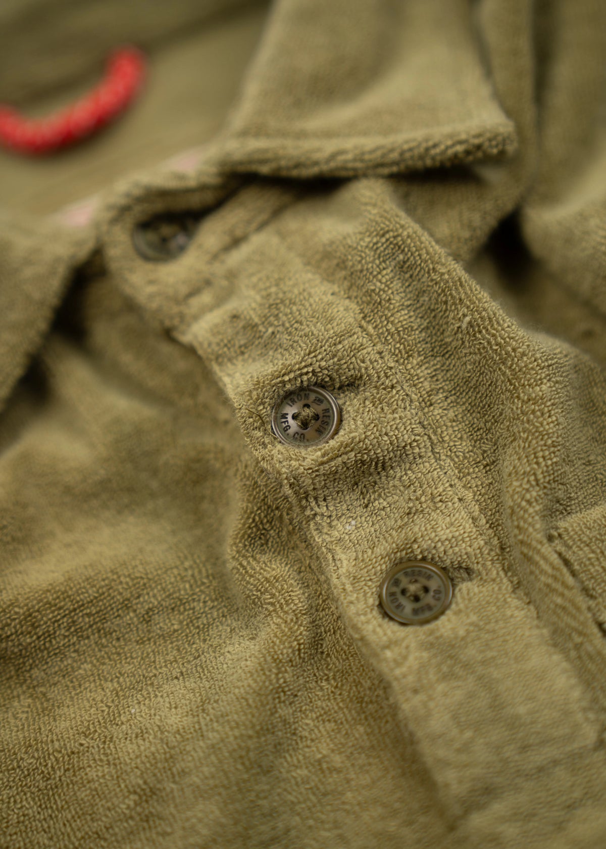 Close-up of a green textured shirt with two visible buttons and a red thread loop, highlighting fabric detail and design.