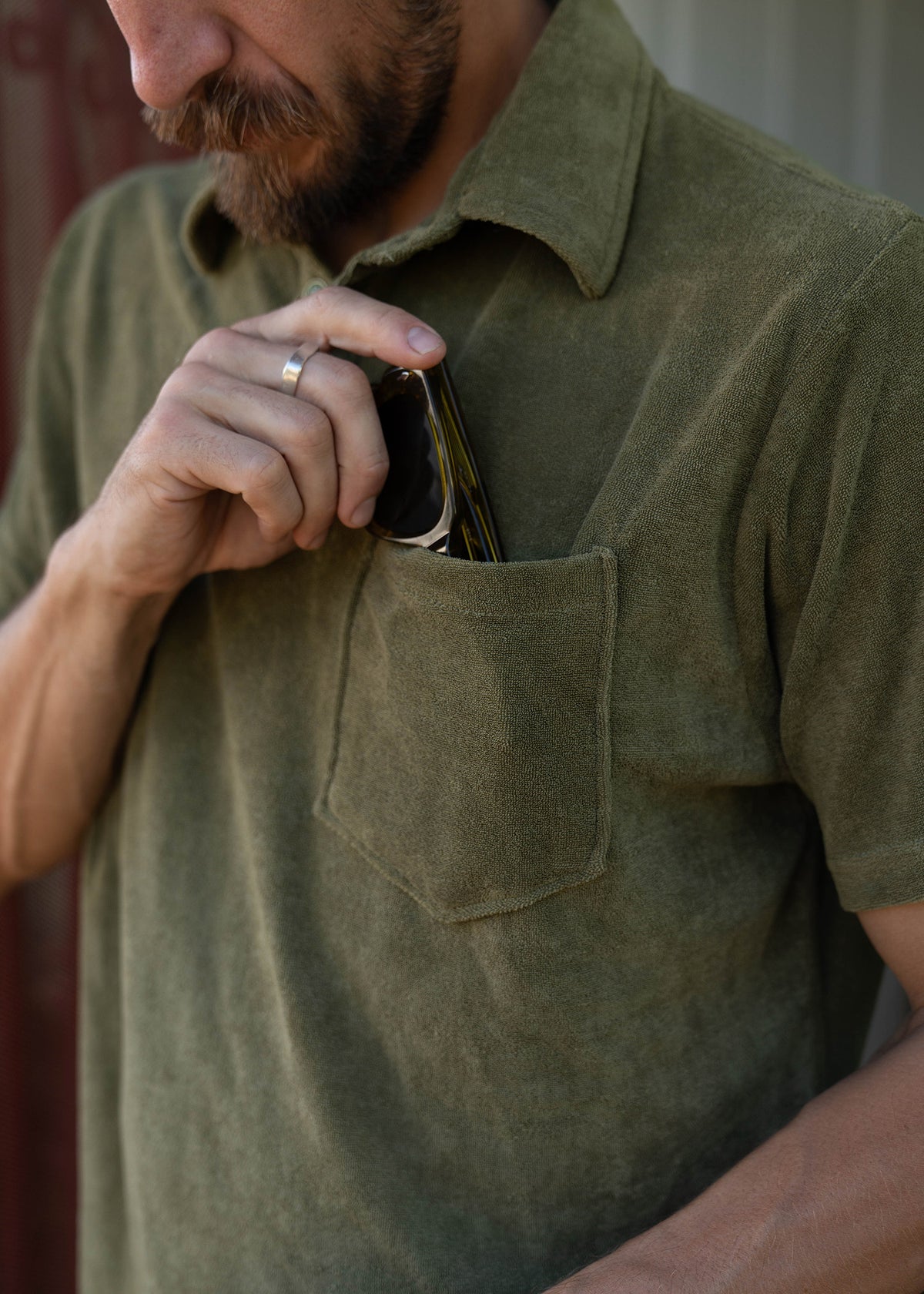 Man in a green shirt places a smartphone into his chest pocket, wearing a silver ring. Close-up shot focuses on the action.