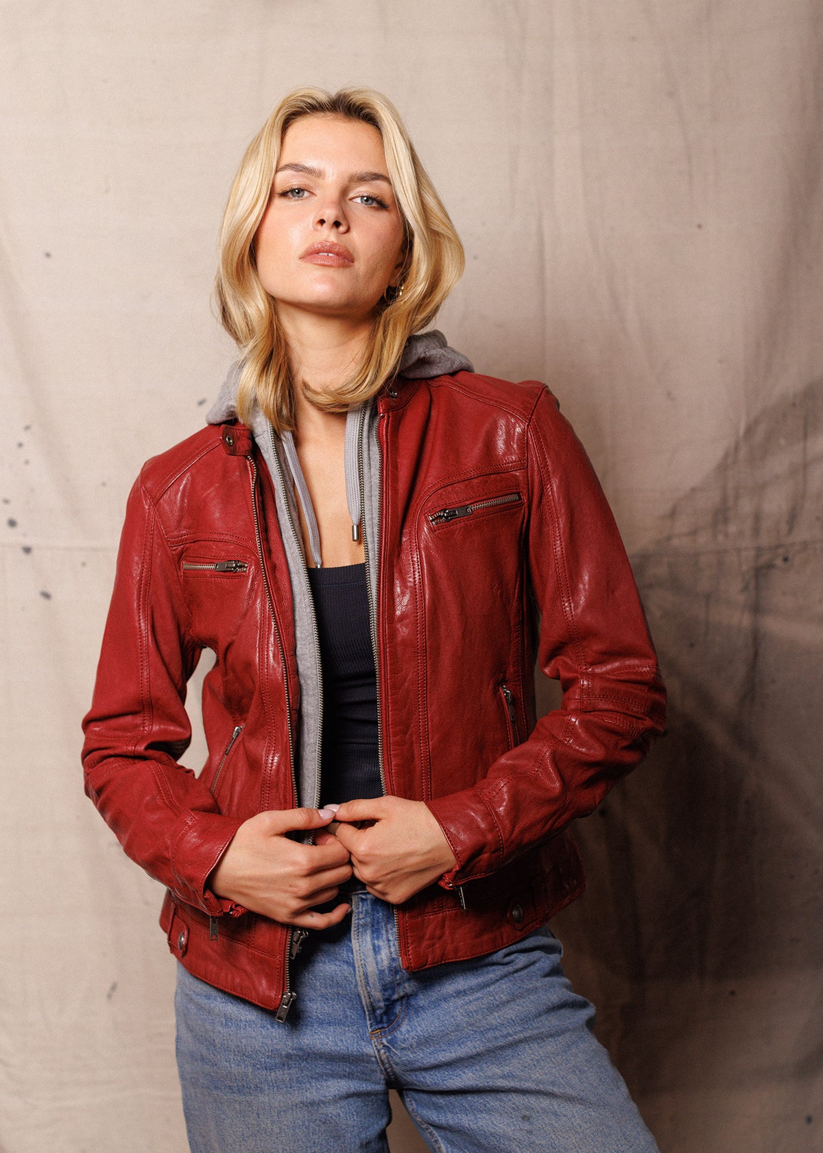 Blonde woman in a red leather jacket and jeans stands confidently against a neutral backdrop, exuding a stylish, casual vibe.