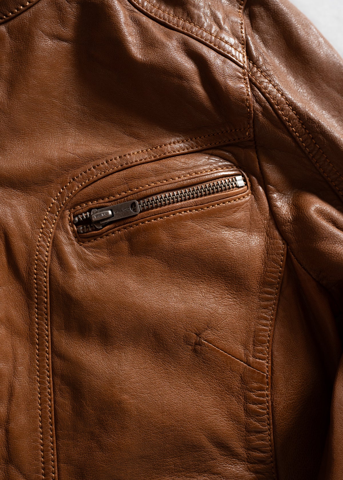Close-up of a brown leather jacket showing detailed stitching and a zippered pocket, highlighting texture and craftsmanship.