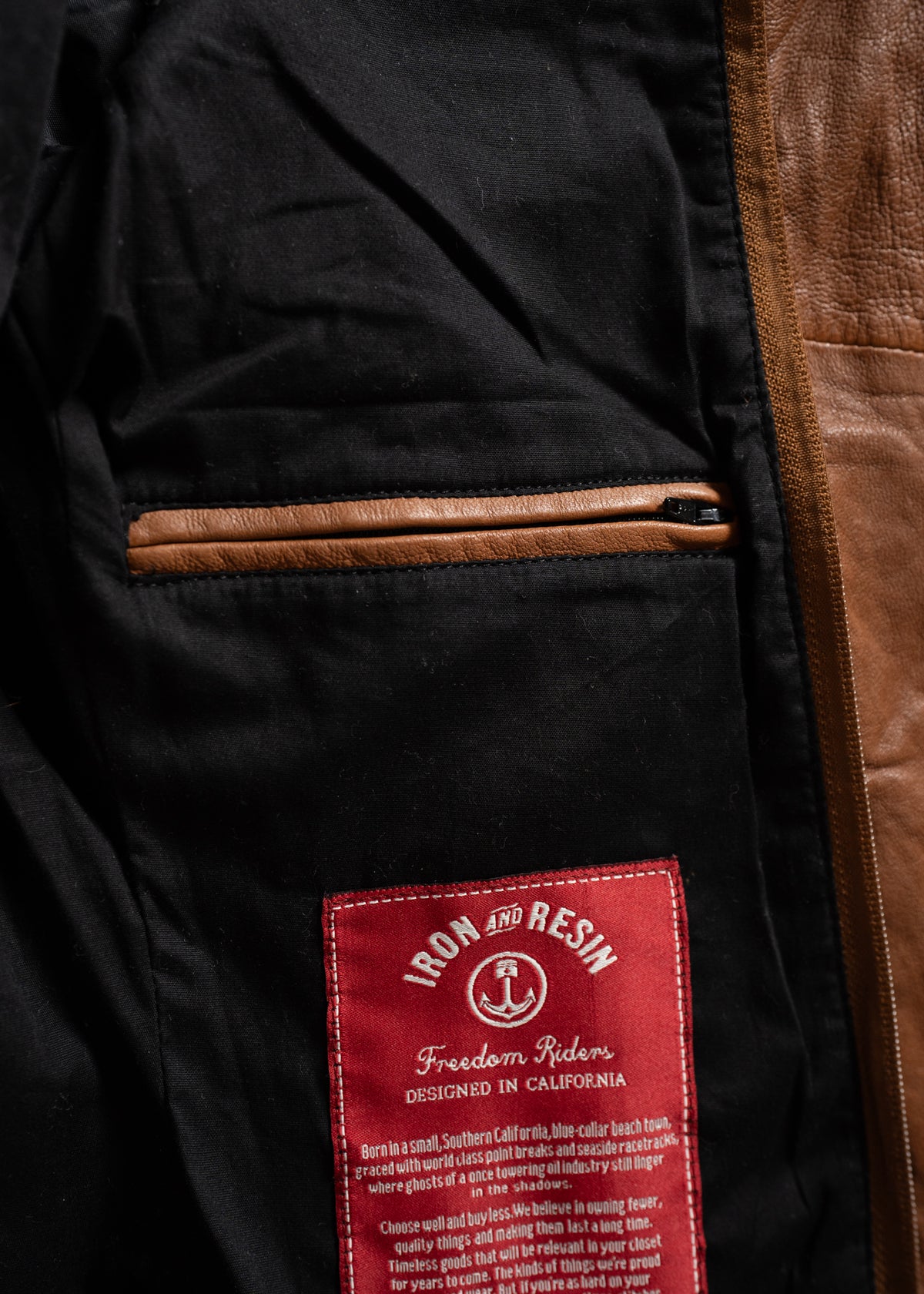 Close-up of a jacket's interior, showing a brown leather pocket and a red "Iron and Resin" label with text about its California design.