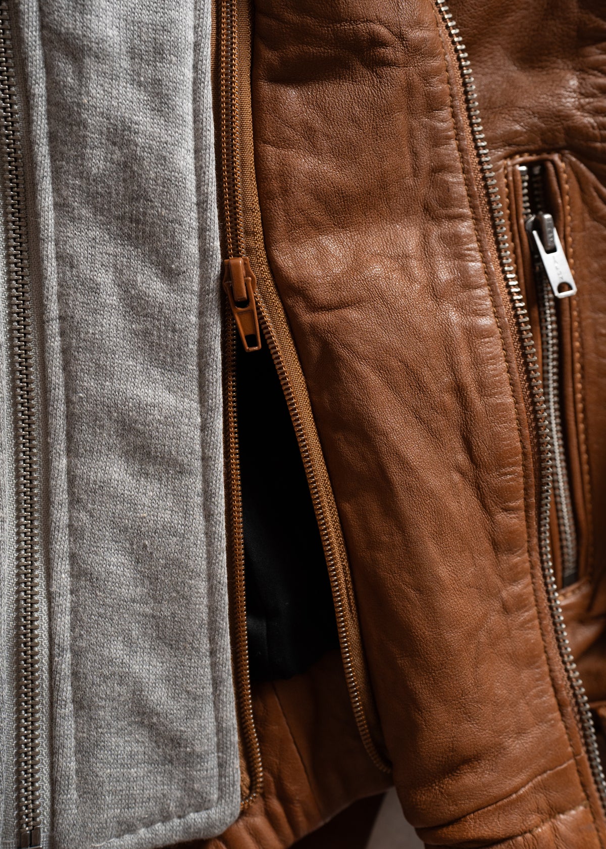 Close-up of a brown leather jacket with visible zippers and a gray fabric lining, highlighting texture and detail.
