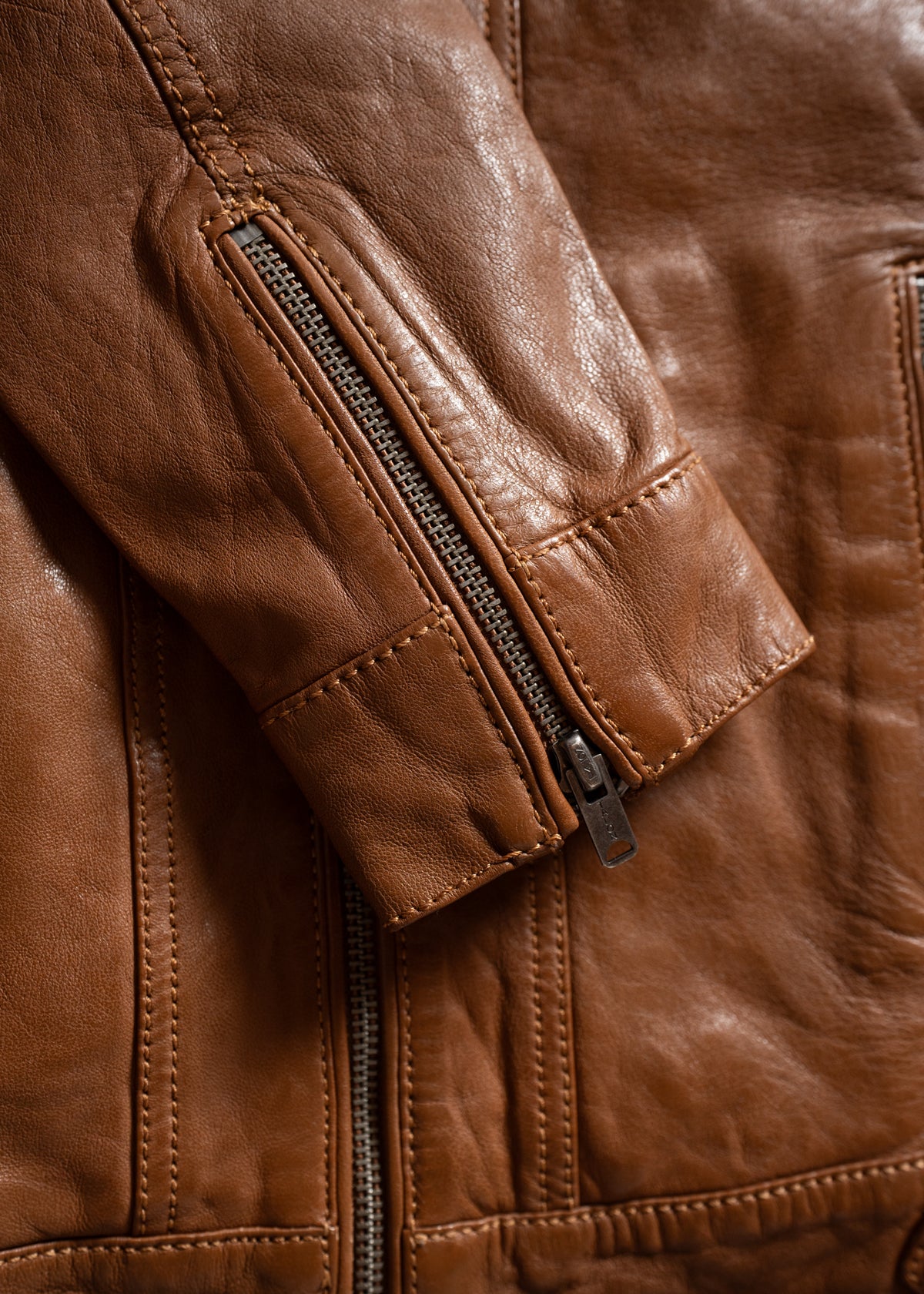 Close-up of a brown leather jacket sleeve with a visible zipper detail, showcasing the texture and stitching of the material.