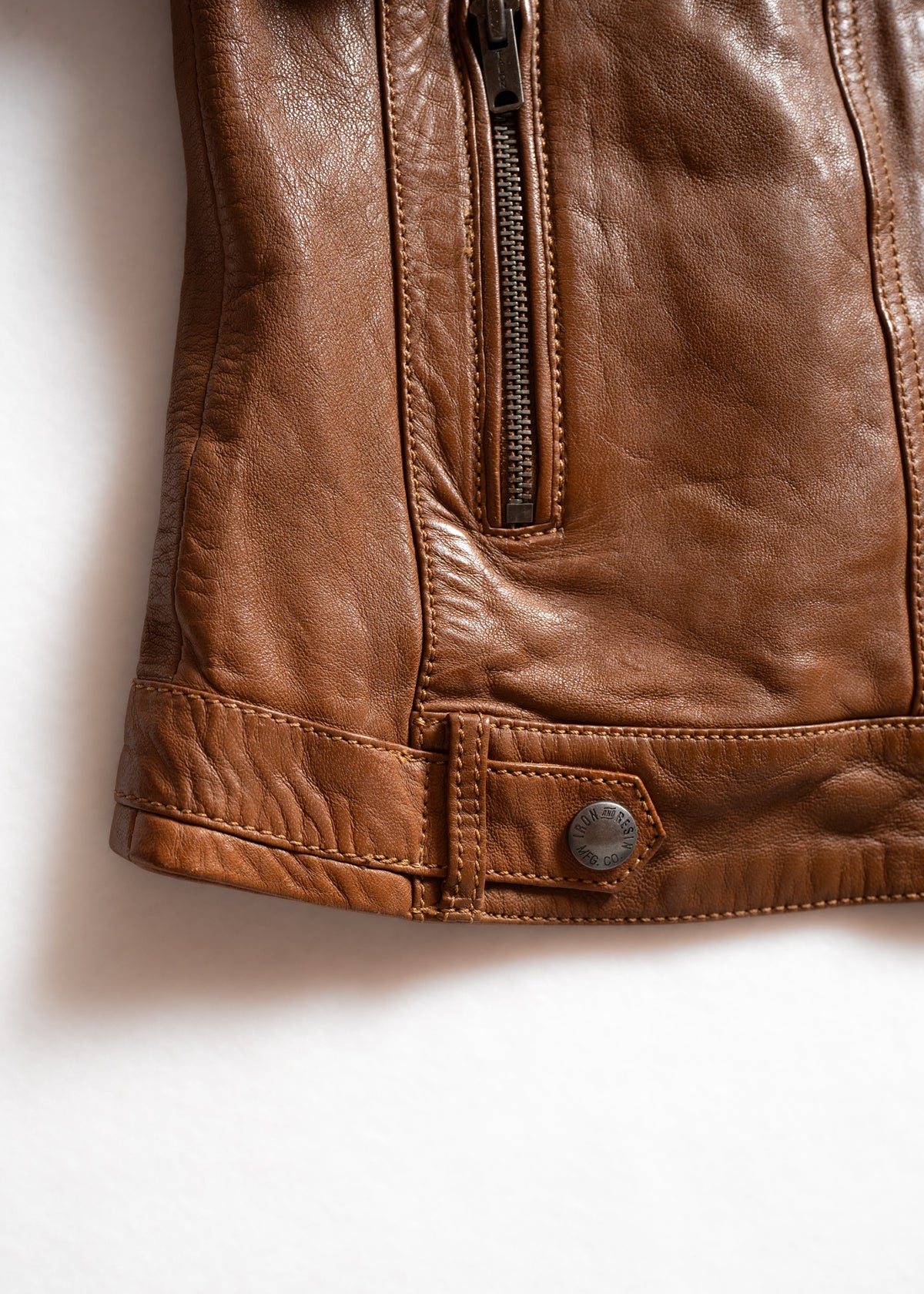 Close-up of a brown leather jacket's zipper and button detail on a white background, highlighting texture and craftsmanship.