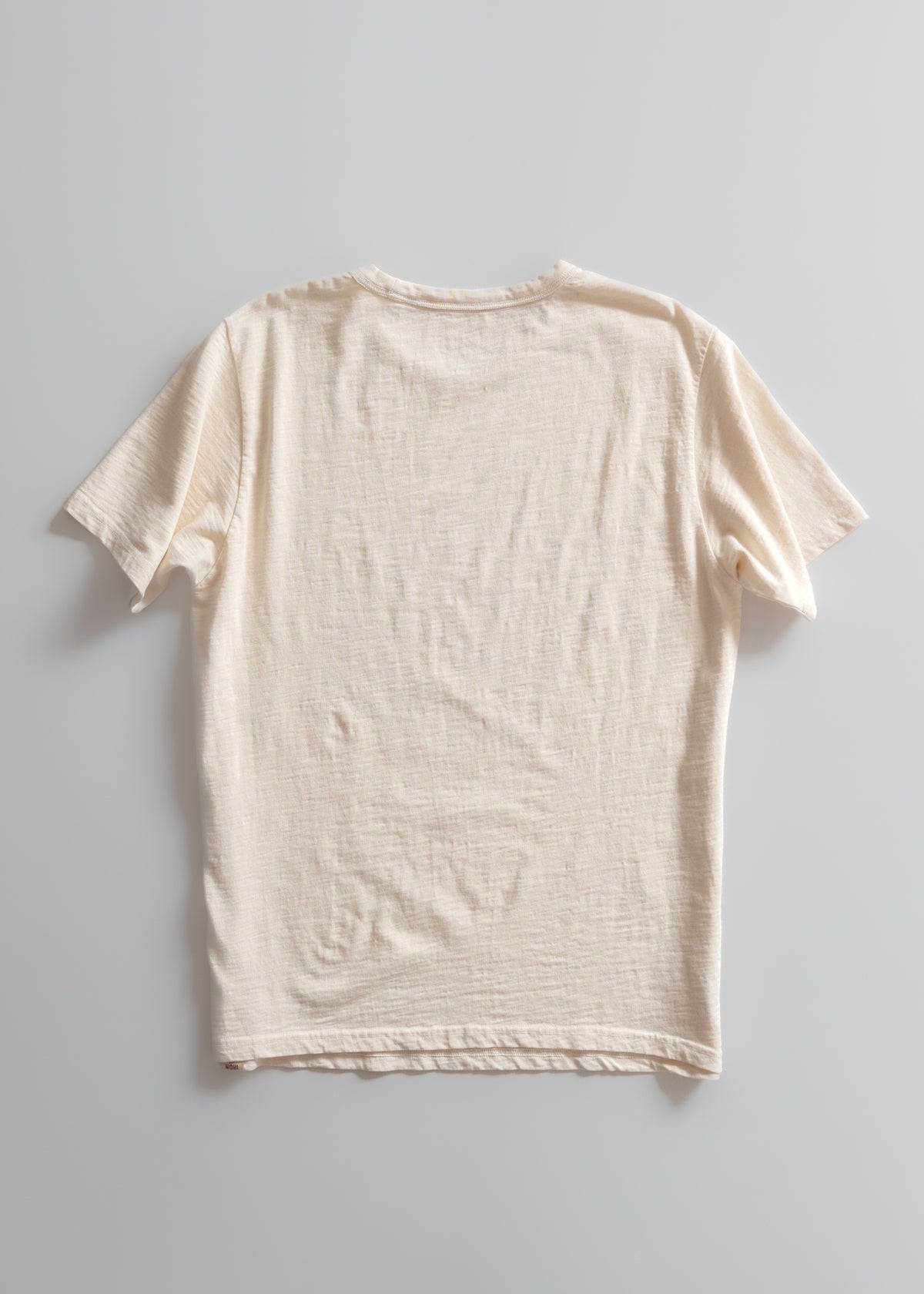A plain beige T-shirt is laid flat on a light gray background, showcasing its texture and short sleeves in a minimalist style.