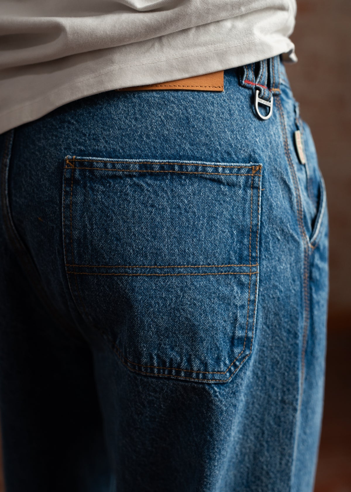 Close-up of blue denim jeans with a visible back pocket, worn with a white shirt. A leather patch and metal loop are visible.