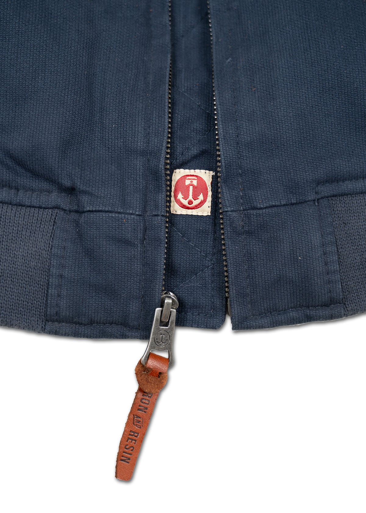 Close-up of a navy jacket zipper with a brown leather pull tab labeled "Iron and Resin" and a red anchor logo on the fabric.