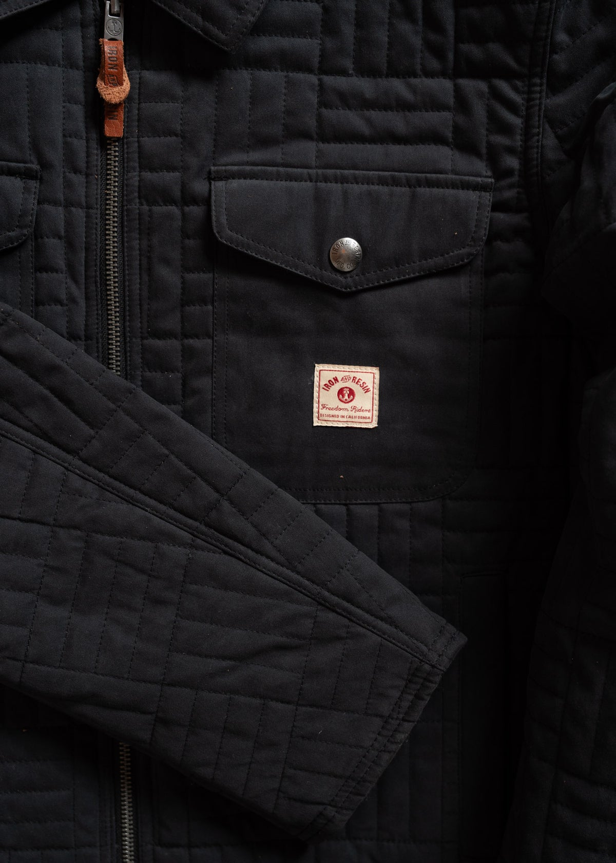 Close-up of a black quilted jacket with a front pocket, metal snap, zipper, and a small red and white "Iron Heart" patch.