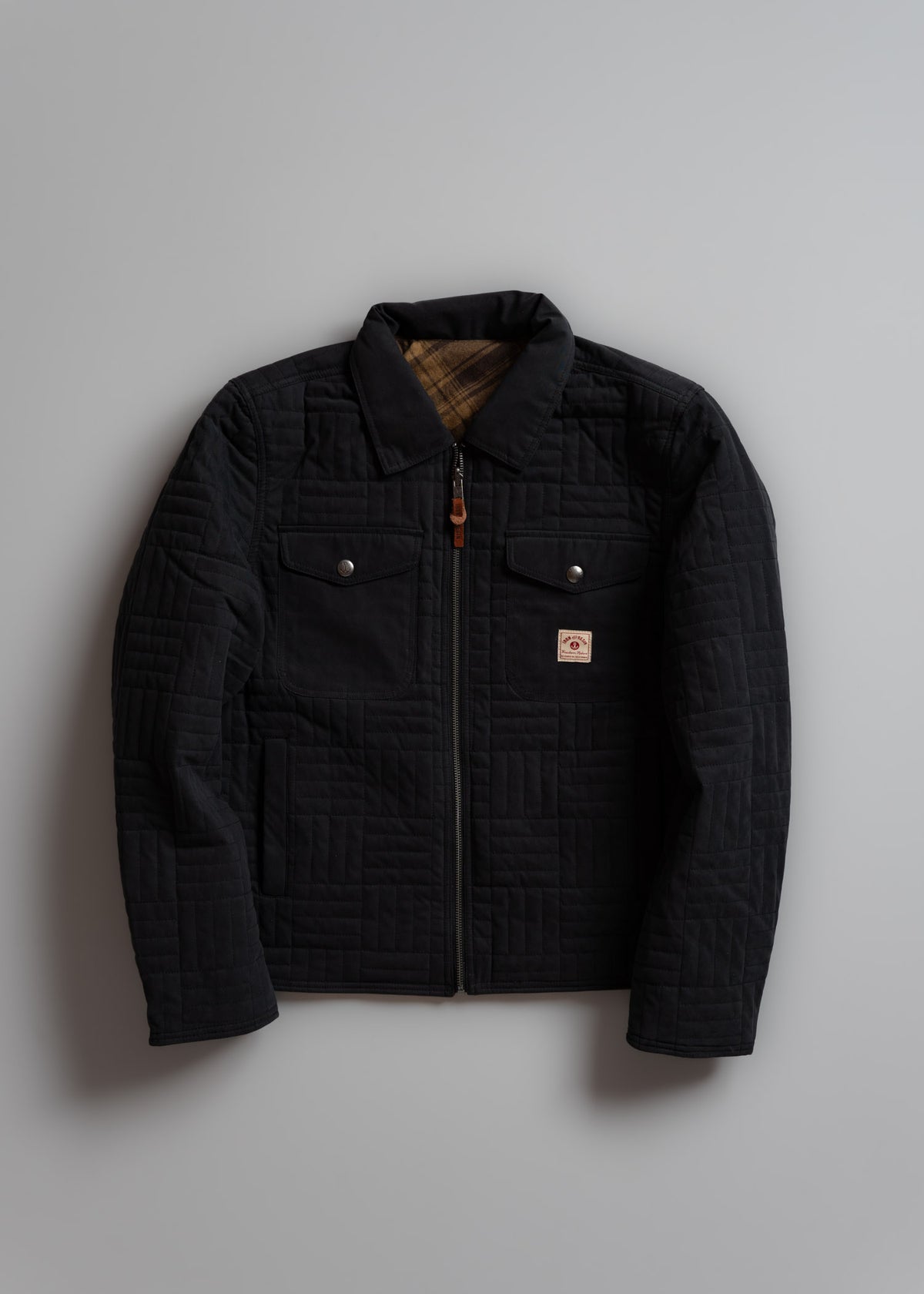 Black quilted jacket with plaid lining, buttoned pockets, and zipper, displayed on a gray background.