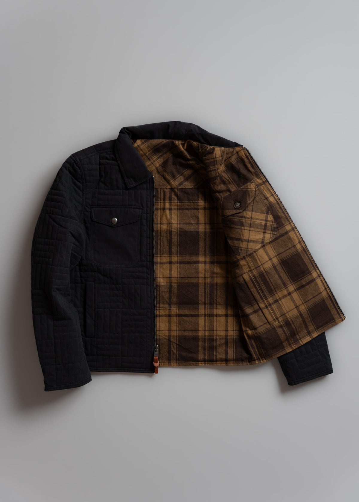 A black quilted jacket with a brown plaid lining is open, showcasing its stylish interior design on a plain background.