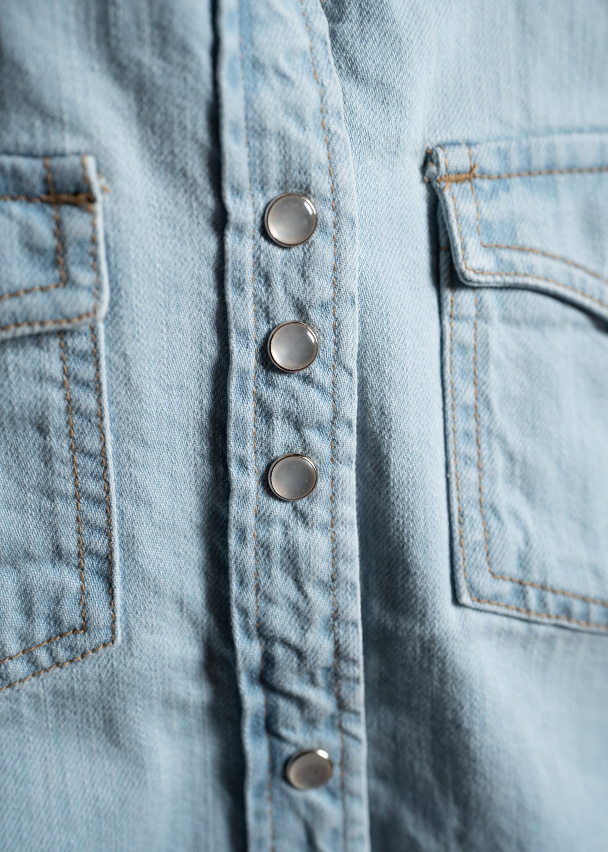Close-up of a light blue denim shirt with metal snap buttons and visible stitching on two pockets, showcasing texture and design.