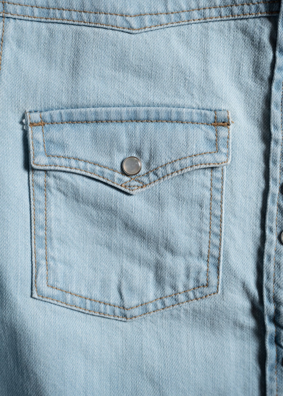 Close-up of a light blue denim shirt pocket with a silver button, showcasing detailed stitching and fabric texture.