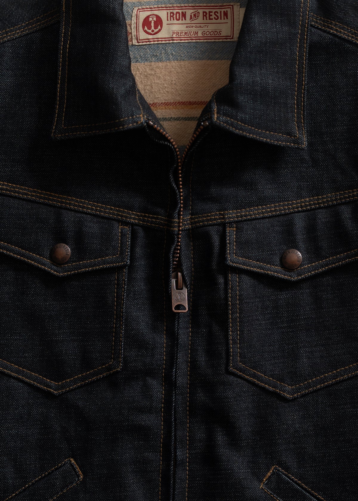 Close-up of a dark denim jacket with a zipper and two chest pockets. Label reads "Iron and Resin Premium Goods."