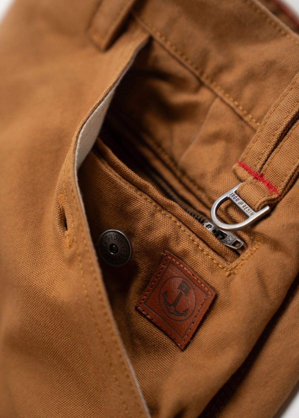 Close-up of brown pants pocket with a metal zipper, button, and leather anchor patch, highlighting rugged, detailed craftsmanship.