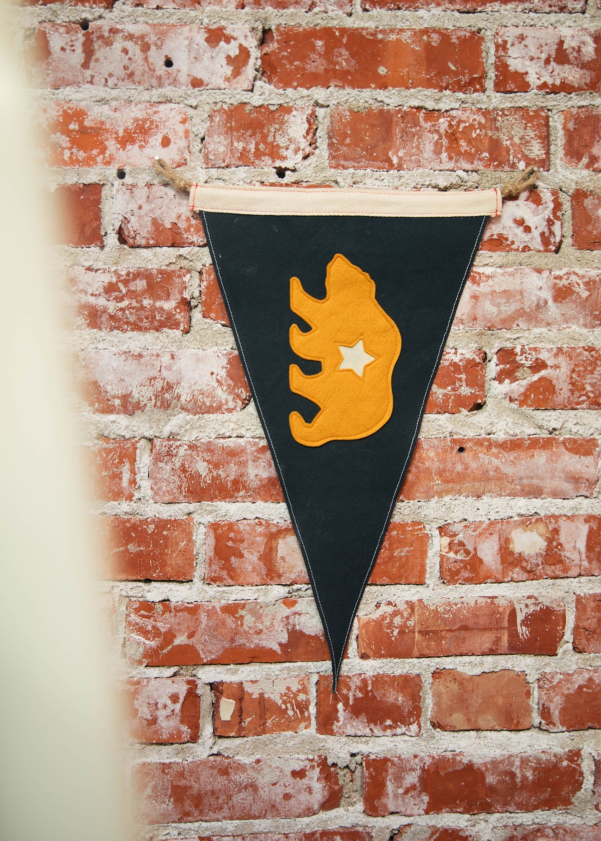 A black pennant with an orange bear and star hangs on a rustic brick wall, adding a touch of whimsy to the textured backdrop.