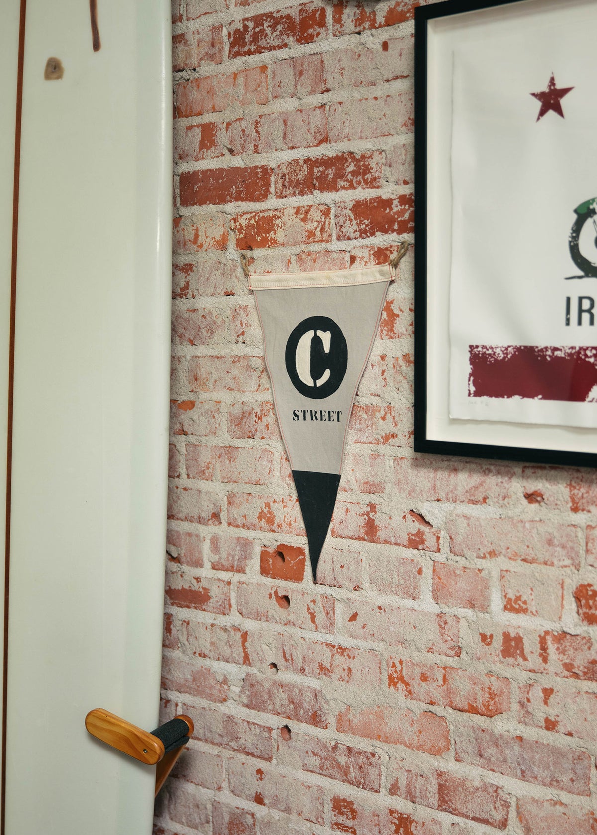 A surfboard leans against a brick wall with a "C Street" pennant and framed art, creating a coastal, vintage vibe.