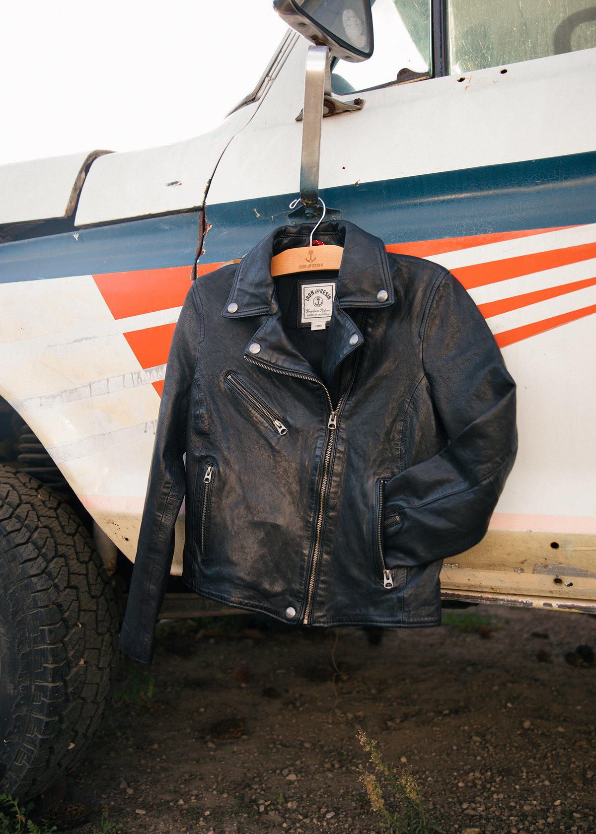 A black leather jacket hangs on a vintage truck's mirror, showcasing its rugged style against the vehicle's retro stripes.