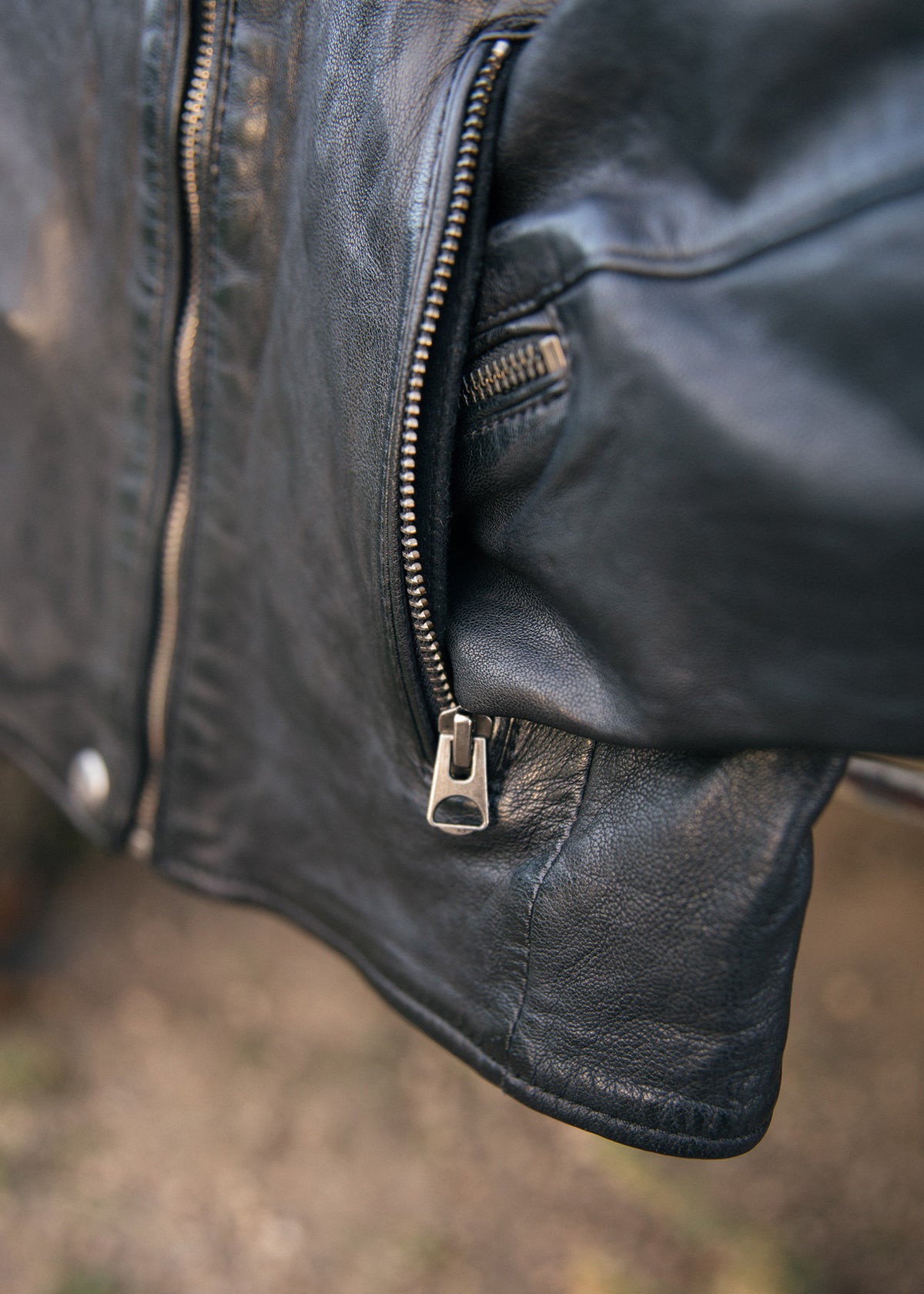 Close-up of a black leather jacket sleeve, highlighting a silver zipper detail against a blurred outdoor background.
