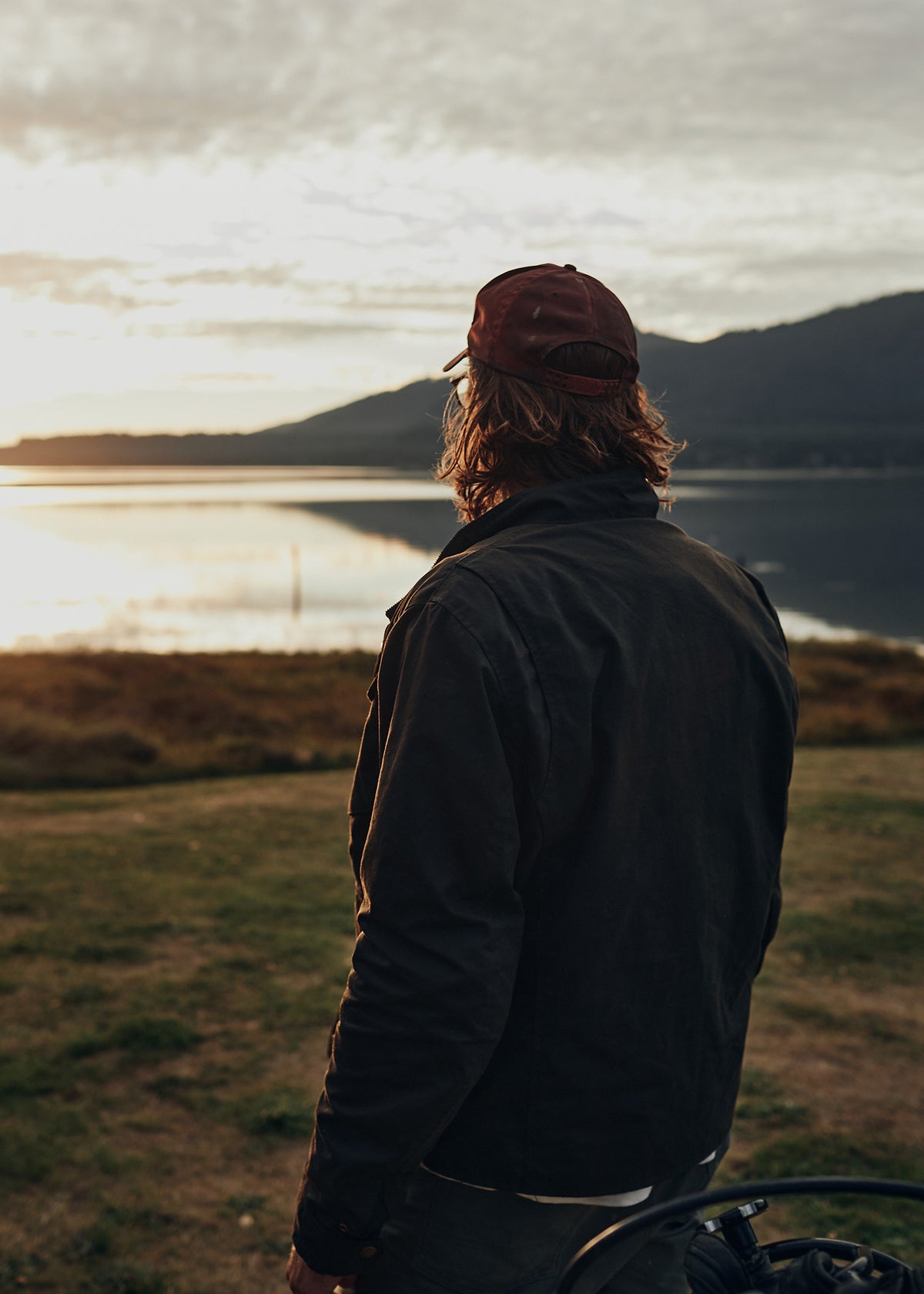 A person in a cap gazes at a serene lake and mountains during sunset, creating a peaceful and reflective scene.