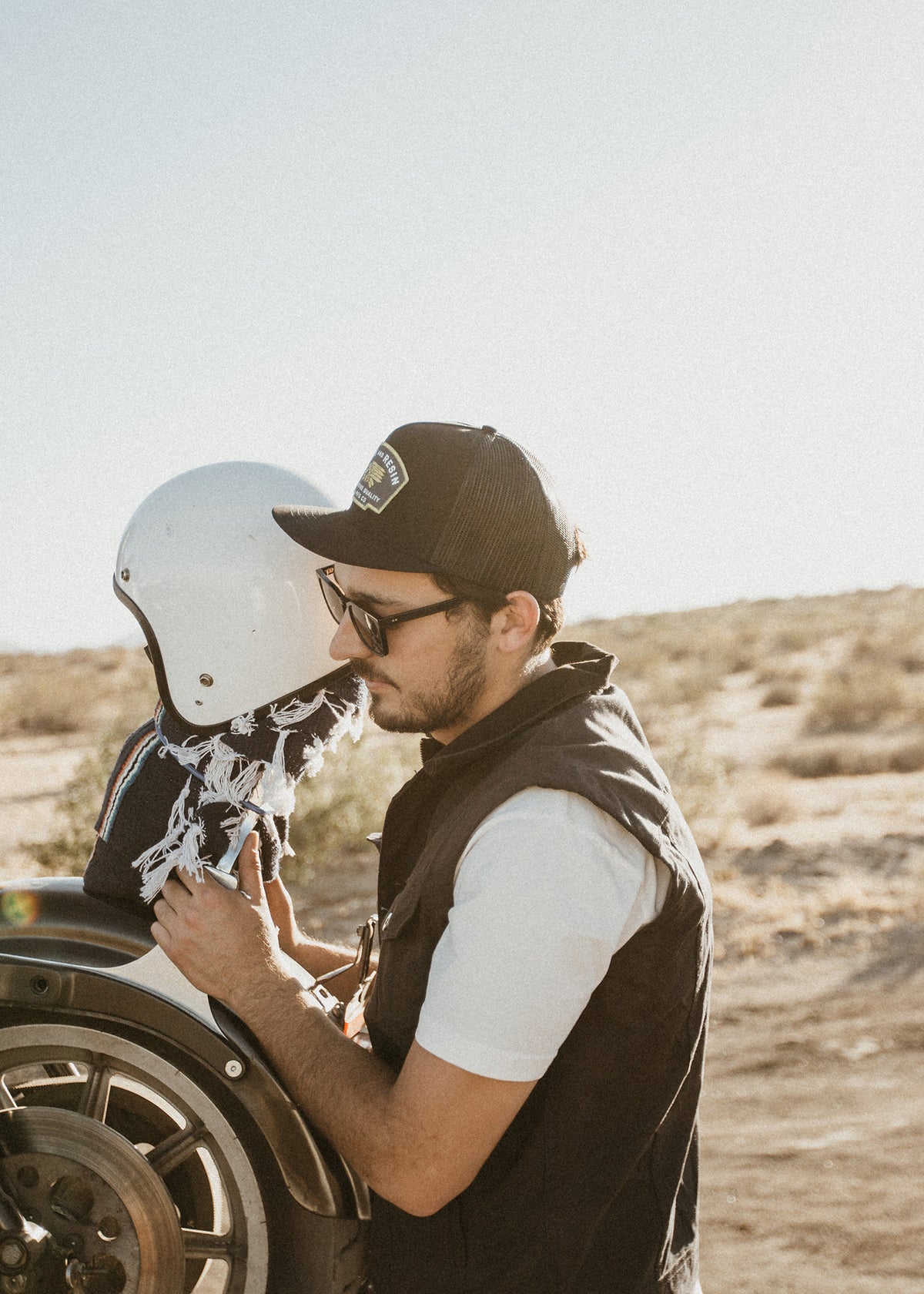 Man in cap and sunglasses adjusts a white helmet on a motorcycle in a sunny desert landscape, creating a serene and adventurous vibe.