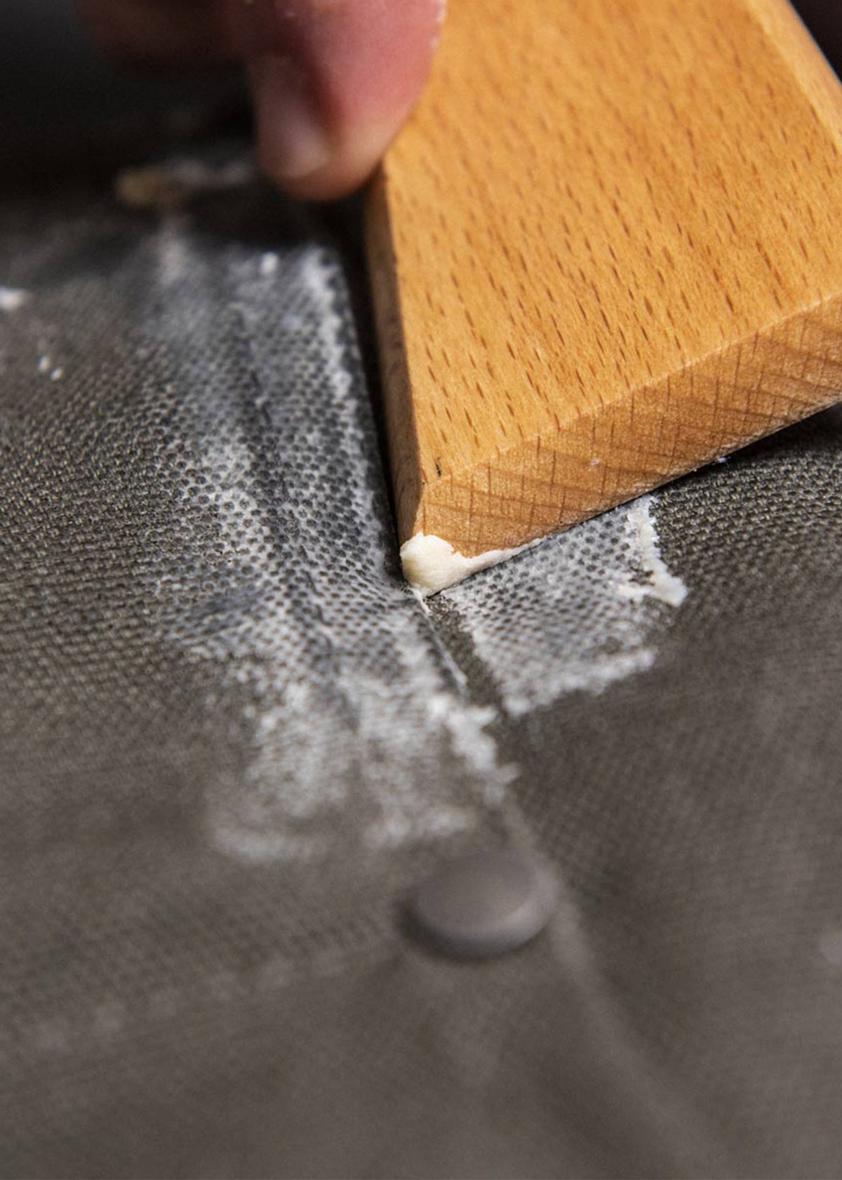 A hand uses a wooden tool to apply white paste on a textured grey surface, focusing on precision and detail in the application.